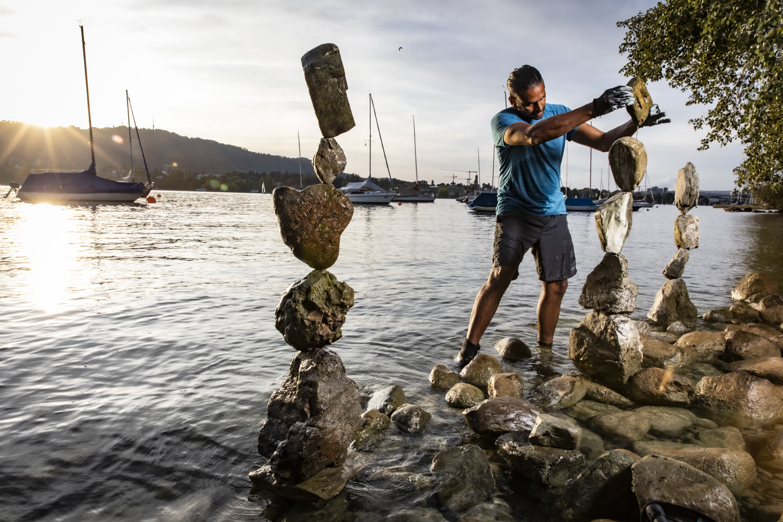 Greg building stone towers in Lake Zürich at golden hour, sailboats behind